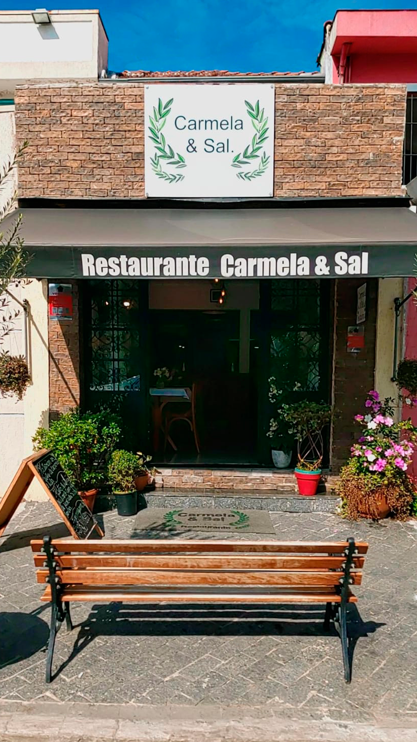 Fachada do restaurante Carmela & Sal com toldo verde, placa de louros e banco de madeira na calçada